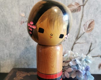 Puede incluir: Muñeca Kokeshi de madera con cabeza redondeada y cuerpo cilíndrico. La cabeza presenta cabello negro pintado, ojos negros pequeños y una boca roja simple. Acentos florales rosas adornan los lados. El cuerpo tiene bandas rojas y negras. Artesanía tradicional japonesa.