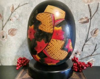May include: A black wooden egg-shaped box with a painted design of a woman in a red and yellow kimono with red maple leaves. The box is sitting on a black wooden base.