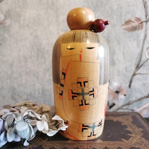 Könnte beinhalten: Eine Kokeshi-Puppe aus Holz mit rundem Körper und Kopf. Die Puppe hat bemalte Details, darunter ein Gesicht, Haare und einen gemusterten Kimono in Orange, Schwarz und Grün. Auf dem Kopf sitzt eine kleine Holzkugel mit einem roten beerenartigen Akzent.