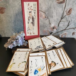 Può includere: Un set di carte d'arte giapponesi con bordi dorati, con illustrazioni e calligrafia. Le carte sono presentate con una scatola rossa, che ha un disegno di una persona. Le carte sono su una superficie scura.