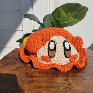 May include: A crocheted plush toy in the shape of a character with orange, white, and brown details. The toy has large eyes and a smiling expression.