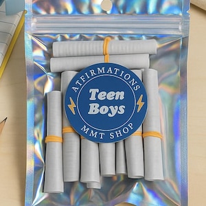 Pode incluir: Uma bolsa de plástico holográfica contendo vários pequenos rolos de papel cinza, apertados e presos com elásticos finos laranja. Um adesivo azul circular na frente apresenta o texto "AFFIRMATIONS Teen Boys MMT SHOP" com dois pequenos ícones de raios amarelos.