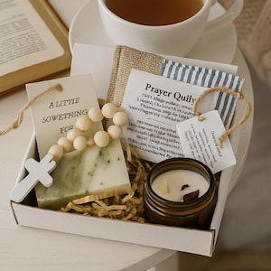 Könnte beinhalten: Eine weiße Geschenkbox mit grünem Seifenstück, Holzrosenkranz, kleiner Kerze und Anhänger mit der Aufschrift "A Little Something For". Eine Tasse Tee und ein offenes Buch stehen daneben. Ruhige, neutrale Farbgebung.