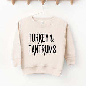 Pode incluir: Camisola bege com o texto preto "TURKEY & TANTRUMS". A camisola est&aacute; pendurada num cabide branco sobre um fundo branco. A camisola tem mangas compridas e gola redonda.