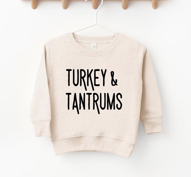 Pode incluir: Camisola bege com o texto preto "TURKEY & TANTRUMS". A camisola est&aacute; pendurada num cabide branco sobre um fundo branco. A camisola tem mangas compridas e gola redonda.