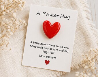 May include: A white rectangular card featuring a glossy red heart-shaped token attached to the center. The text reads A Pocket Hug, A little heart from me to you, filled with lots of love and big hugs too! Love you lots, with a small red heart icon.