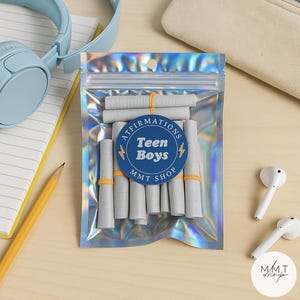May include: A clear, iridescent pouch containing rolled-up white paper scrolls secured with orange bands. A blue and white circular label reads "Affirmations Teen Boys MMT Shop." The background includes a light blue headset, a pencil, and white earbuds.
