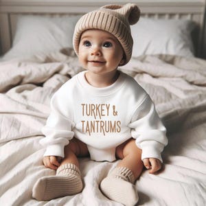 Pode incluir: Um beb&eacute; sorridente a usar um gorro de malha bege, um camisola branca com o texto "TURKEY & TANTRUMS" e meias bege. O beb&eacute; est&aacute; sentado numa cama com um edred&atilde;o de tons neutros.