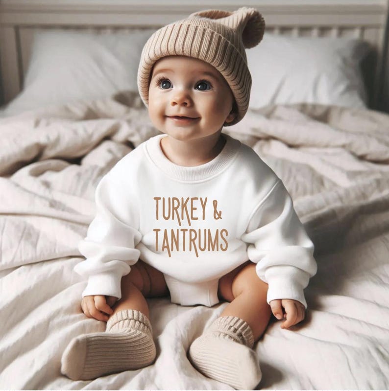Pode incluir: Um beb&eacute; sorridente a usar um gorro de malha bege, um camisola branca com o texto "TURKEY & TANTRUMS" e meias bege. O beb&eacute; est&aacute; sentado numa cama com um edred&atilde;o de tons neutros.