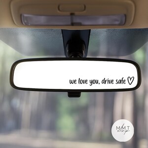 May include: A car's interior rearview mirror displaying the message "we love you, drive safe" in black script, with a heart symbol. The background is a blurred view of a forest, with the car's roof and windscreen visible.