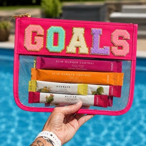 Puede incluir: Un estuche rosa brillante con cremallera y panel frontal transparente que muestra la palabra GOALS en letras coloridas y texturizadas de estilo universitario. Dentro hay cuatro sobres rectangulares de suplementos dietéticos etiquetados como Slim Hunger Control, Hydrate y Active.