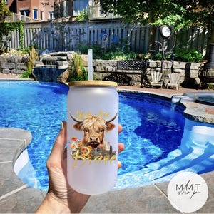 May include: A frosted glass tumbler with a bamboo lid and a clear straw. The tumbler features a graphic of a Highland cow with orange flowers and the gold text "You Are Brave" printed on the front surface of the glass container.
