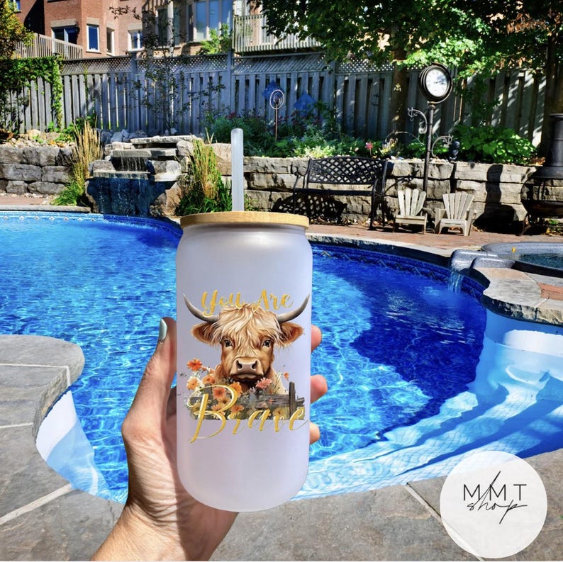 May include: A frosted glass tumbler with a bamboo lid and a clear straw. The tumbler features a graphic of a Highland cow with orange flowers and the gold text "You Are Brave" printed on the front surface of the glass container.