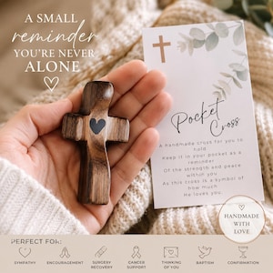 May include: A small dark brown wooden pocket cross with a black heart icon in the center. Beside it is a white card with the text Pocket Cross, a small cross symbol, and eucalyptus illustrations, along with the text A small reminder you're never alone.