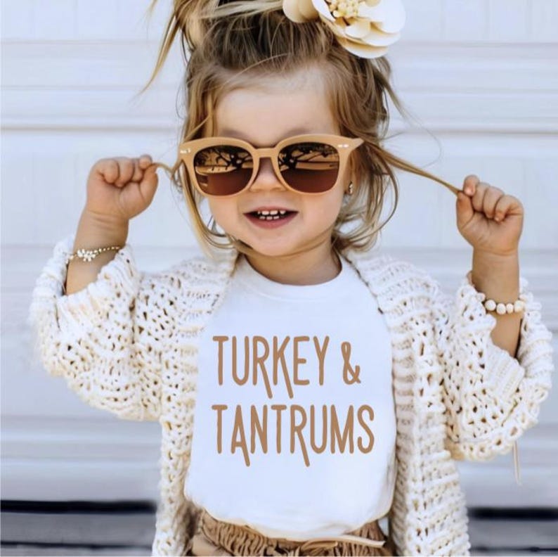 Pode incluir: Uma crian&ccedil;a pequena usando &oacute;culos de sol e uma camiseta branca com as palavras "TURKEY & TANTRUMS" em marrom. A crian&ccedil;a tamb&eacute;m est&aacute; usando um cardig&atilde; de malha aberta creme e uma pulseira. Um acess&oacute;rio de cabelo de flor &eacute; vis&iacute;vel.