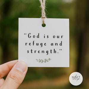 May include: A square white card with the quote "God is our refuge and strength." in black text. The card is suspended by a natural twine string. A small, circular logo with the text "MMT shop" is in the bottom right corner.