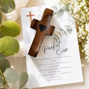 May include: A dark brown wooden pocket cross featuring a small black heart in the center, resting on a white card printed with the text Pocket Cross and a poem, surrounded by green eucalyptus leaves and small white flowers.