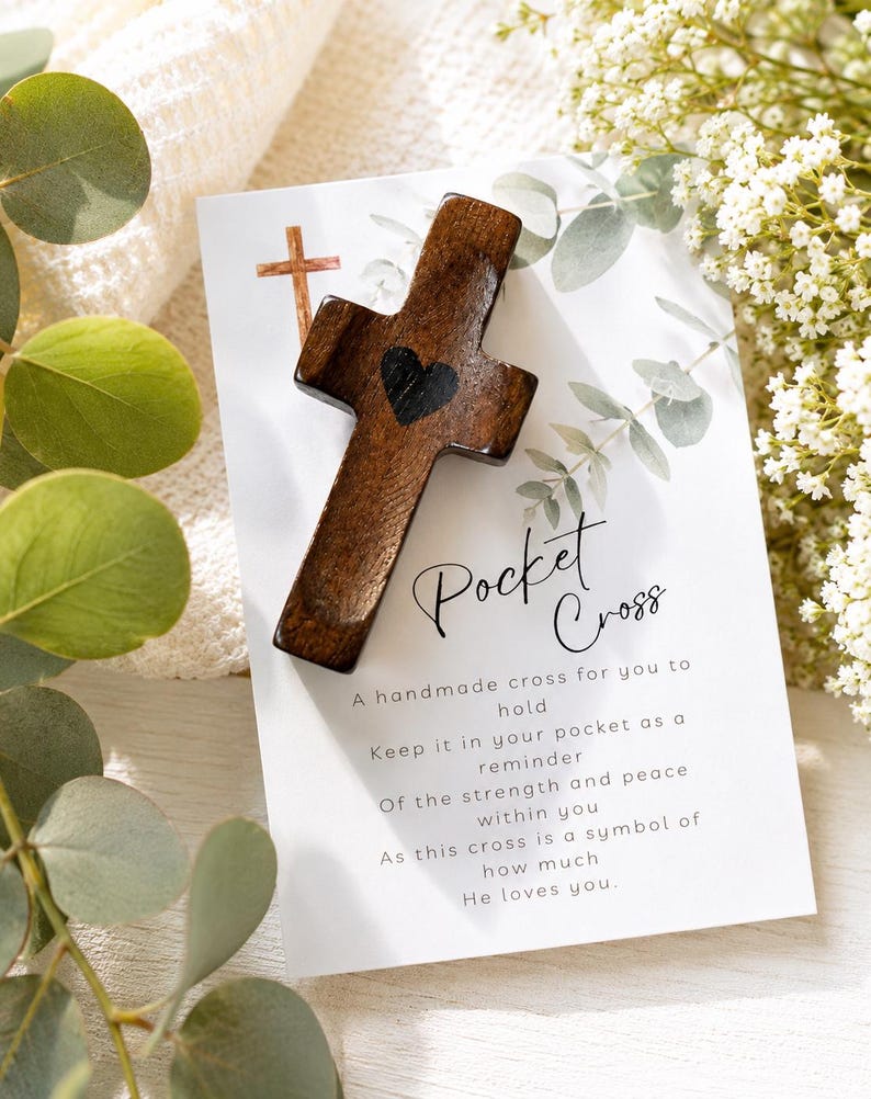 May include: A dark brown wooden pocket cross featuring a small black heart in the center, resting on a white card printed with the text Pocket Cross and a poem, surrounded by green eucalyptus leaves and small white flowers.