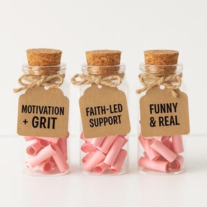 May include: Three glass bottles with cork stoppers and attached tags. The tags read "MOTIVATION + GRIT", "FAITH-LED SUPPORT", and "FUNNY & REAL". Each bottle contains pink rolled paper. The bottles are tied with twine.