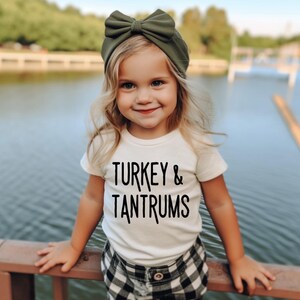 Pode incluir: Uma crian&ccedil;a vestindo uma camiseta branca com o texto preto "TURKEY & TANTRUMS". A crian&ccedil;a tamb&eacute;m est&aacute; usando uma faixa de cabe&ccedil;a verde com um la&ccedil;o grande e shorts xadrez preto e branco. O fundo apresenta um corpo de &aacute;gua.