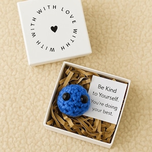 May include: A blue crocheted ball with black eyes sits in a white box filled with brown paper. A card inside the box reads "Be Kind to Yourself. You're doing your best." The box is inside a larger white box with "WITH LOVE" and a heart.
