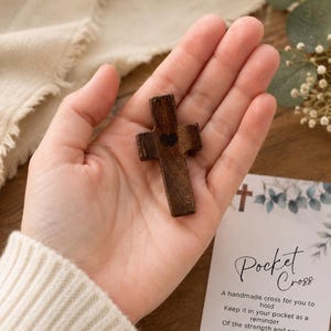May include: A small, dark brown wooden cross featuring a tiny black heart symbol at its center. The cross is accompanied by a white card with the text Pocket Cross and a poem about strength and peace, decorated with a small cross and leaf illustration.