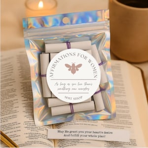 May include: A holographic pouch filled with rolled paper scrolls, secured with purple twine. A circular label reads "AFFIRMATIONS FOR WOMEN" with a bee graphic and the text "As long as you live there's something new everyday" and "MMT SHOP".