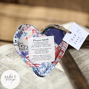 May include: A heart-shaped prayer quilt made of various patterned fabrics, with a tag that reads "God is love." The quilt has a sewn-on label with a poem. A jute twine is attached for hanging. The background includes an open book.