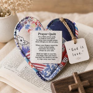 Op de afbeelding: Een hartvormig stoffen kussentje met een patchworkpatroon van blauwe, rode en witte bloemen- en streepstoffen. Een wit vierkant lapje met zwarte tekst is in het midden vastgenaaid. Een klein vierkant label aan een touwtje bevat de tekst God is love.