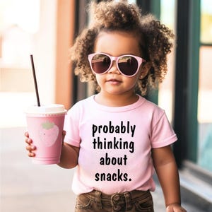 Pode incluir: Uma criança usando óculos de sol rosa e uma camiseta rosa que diz "probably thinking about snacks". A criança está segurando uma xícara rosa com um desenho de morango e um canudo.