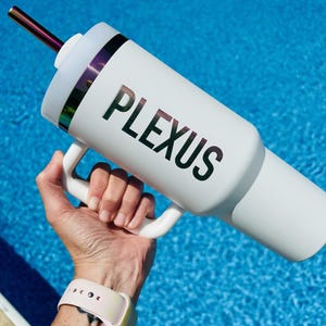 Puede incluir: Vaso blanco con asa y pajita. La palabra "PLEXUS" está impresa en negro en el lateral. La pajita tiene un acabado metálico. El vaso se sostiene sobre un fondo de agua azul.