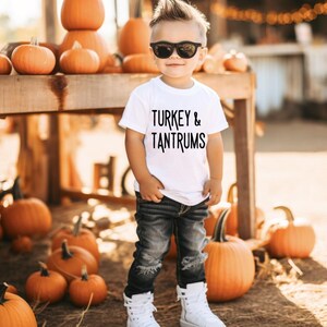Pode incluir: Uma crian&ccedil;a usando &oacute;culos de sol, uma camiseta branca com a frase "TURKEY & TANTRUMS", jeans pretos rasgados e t&ecirc;nis brancos de cano alto. O fundo apresenta ab&oacute;boras e uma estrutura de madeira, sugerindo um tema de outono.