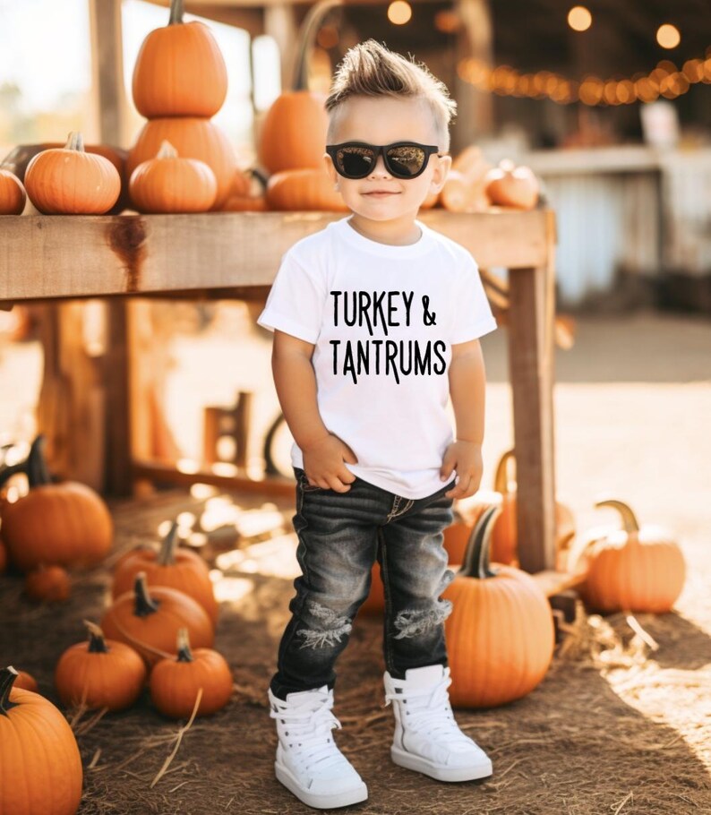 Pode incluir: Uma crian&ccedil;a usando &oacute;culos de sol, uma camiseta branca com a frase "TURKEY & TANTRUMS", jeans pretos rasgados e t&ecirc;nis brancos de cano alto. O fundo apresenta ab&oacute;boras e uma estrutura de madeira, sugerindo um tema de outono.