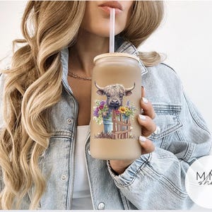 May include: A frosted glass tumbler with a bamboo lid and a clear straw, featuring a colorful graphic of a Highland cow peeking over a wooden fence surrounded by a bouquet of purple, yellow, and green wildflowers against a light background.