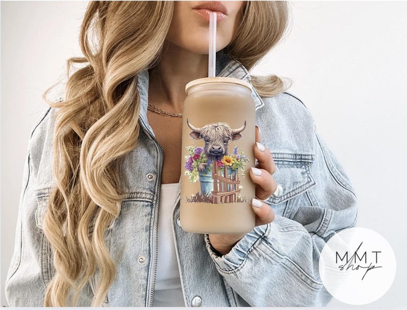 May include: A frosted glass tumbler with a bamboo lid and a clear straw, featuring a colorful graphic of a Highland cow peeking over a wooden fence surrounded by a bouquet of purple, yellow, and green wildflowers against a light background.