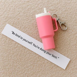May include: A pink tumbler keychain with a white lid and straw, and a silver-colored clip. A white paper strip with the text "Be kind to yourself. You're doing your best." is in the foreground.