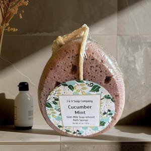 May include: A Cucumber Mint goat milk soap infused bath sponge from J & V Soap Company. The sponge is a light purple color and is packaged in clear plastic. A white bottle with a black cap is to the left of the sponge.