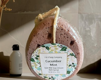 May include: A Cucumber Mint goat milk soap infused bath sponge from J & V Soap Company. The sponge is a light purple color and is packaged in clear plastic. A white bottle with a black cap is to the left of the sponge.