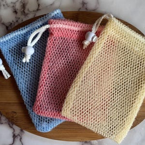 May include: Three mesh soap bags in blue, pink, and yellow, each with a drawstring closure and a white bead. The bags are arranged on a wooden cutting board.