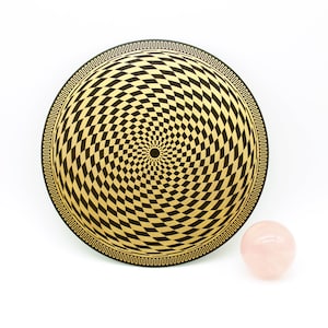 Op de afbeelding: Een zwart en gouden rond bord met een draaiend geometrisch patroon. Een roze rozenkwarts bol ligt op een wit oppervlak naast het bord.