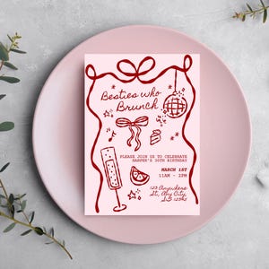 May include: A pink invitation card with the text "Besties who Brunch" in a handwritten style, with illustrations of a disco ball, champagne flute, and cake. The invitation is on a pink plate with a gold fork and white napkin.