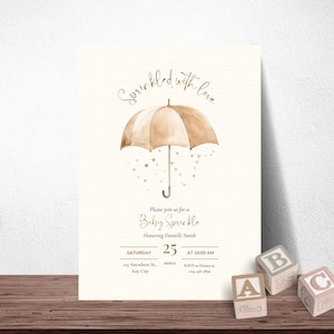 May include: A baby sprinkle invitation with a watercolor umbrella design in shades of brown. The text reads "Sprinkled with love" and includes event details: date, time, and location. Wooden alphabet blocks are in the foreground.