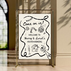 May include: A gold metal frame holds a white sign with black handwritten text and illustrations. The sign reads "Come on in!" and "Welcome to Harry & Sarah's Housewarming."