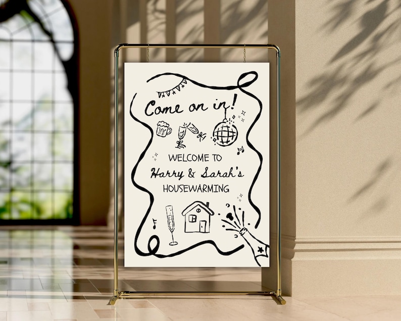 May include: A gold metal frame holds a white sign with black handwritten text and illustrations. The sign reads "Come on in!" and "Welcome to Harry & Sarah's Housewarming."