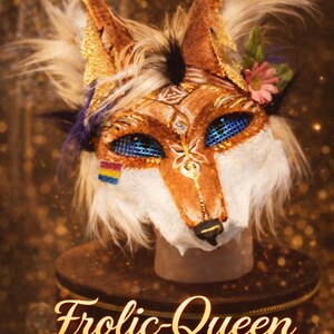 May include: A detailed fox mask with brown, gold, and white fur, adorned with blue eyes and floral accents. The mask features a rainbow flag and the text "Frolic-Queen" in gold script, displayed on a decorative stand.