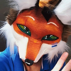 May include: A fox mask with orange, white, and brown fur. The mask has green eyes with black crosshatch details, a black nose, and white accents. The ears are black with white fur tips. The mask is worn over a blue shirt.