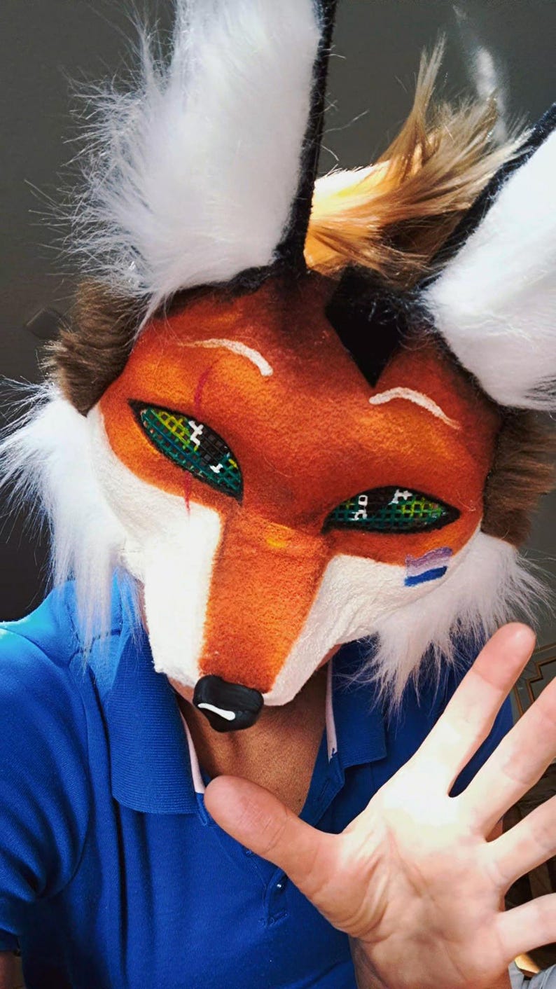 May include: A fox mask with orange, white, and brown fur. The mask has green eyes with black crosshatch details, a black nose, and white accents. The ears are black with white fur tips. The mask is worn over a blue shirt.