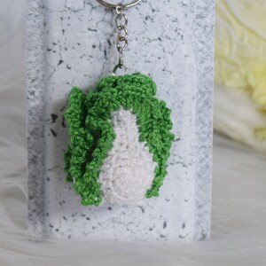 May include: A green and white crocheted keychain shaped like a head of napa cabbage. The keychain is attached to a silver key ring.