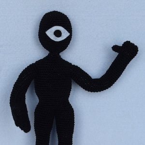 May include: A black crocheted figure with a single white eye. The figure has a human-like form with arms and legs.