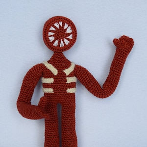 May include: A crocheted red figure with a white circle design on its head. The figure has a white striped pattern on its torso and is standing with one arm raised.